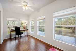 Ein Zimmer mit großen Fenstern, einem Deckenventilator, einem Holzboden und einem Schreibtisch mit Stuhl.