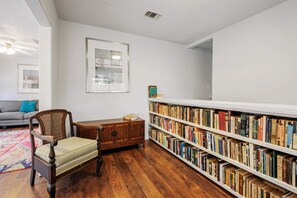 Ein Zimmer mit Holzboden, einem Bücherregal voller Bücher, einem Holzschrank, einem Stuhl und einem Fenster.