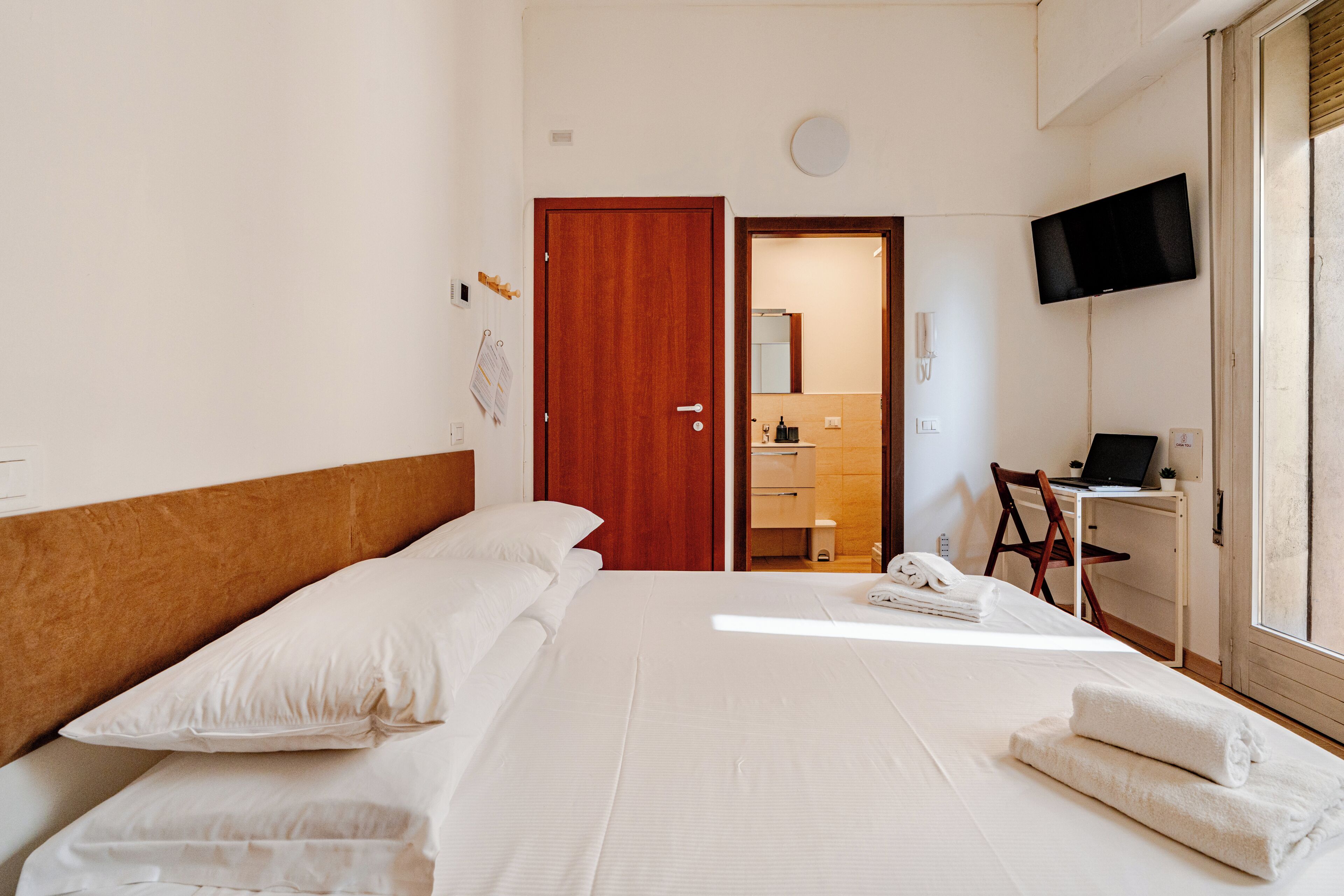 Een hotelkamer met een bed, een bureau met een laptop, een televisie en een badkamer door een open deur zichtbaar.