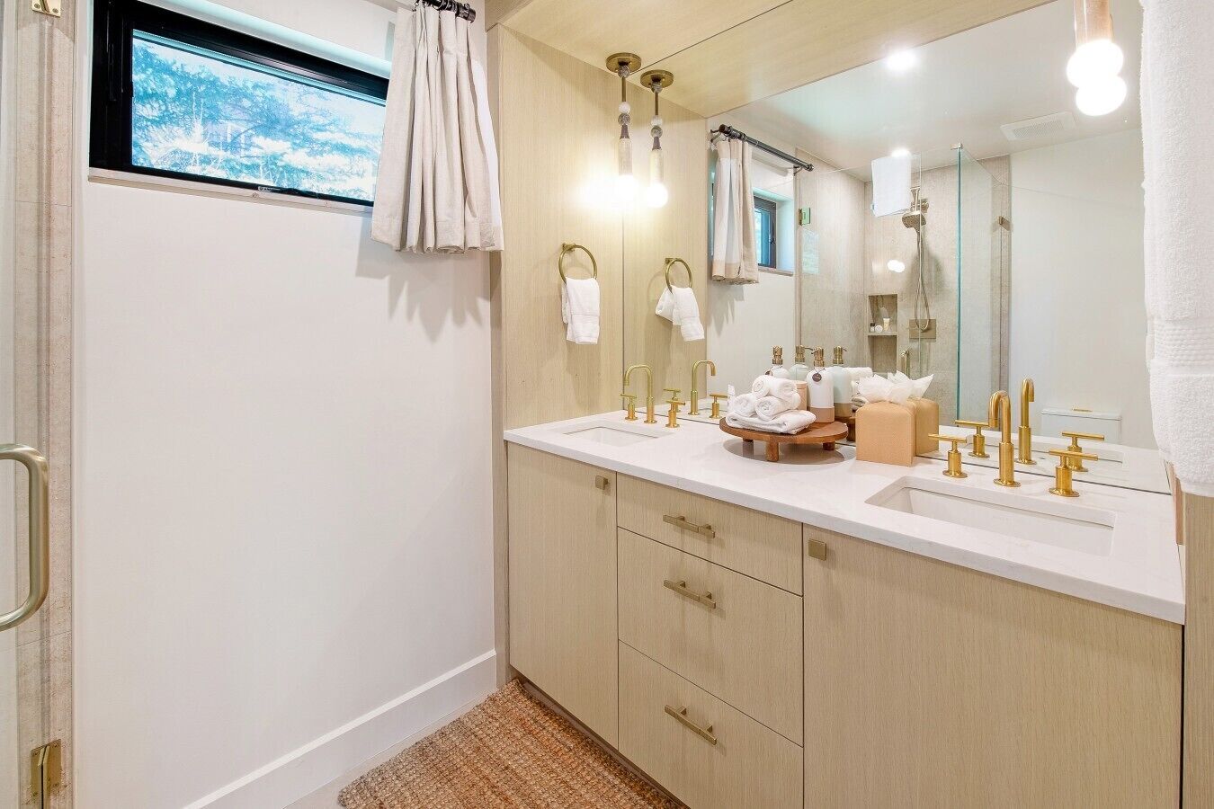 Un baño moderno con un espejo grande, grifos dorados y un ventanal con vistas a árboles.