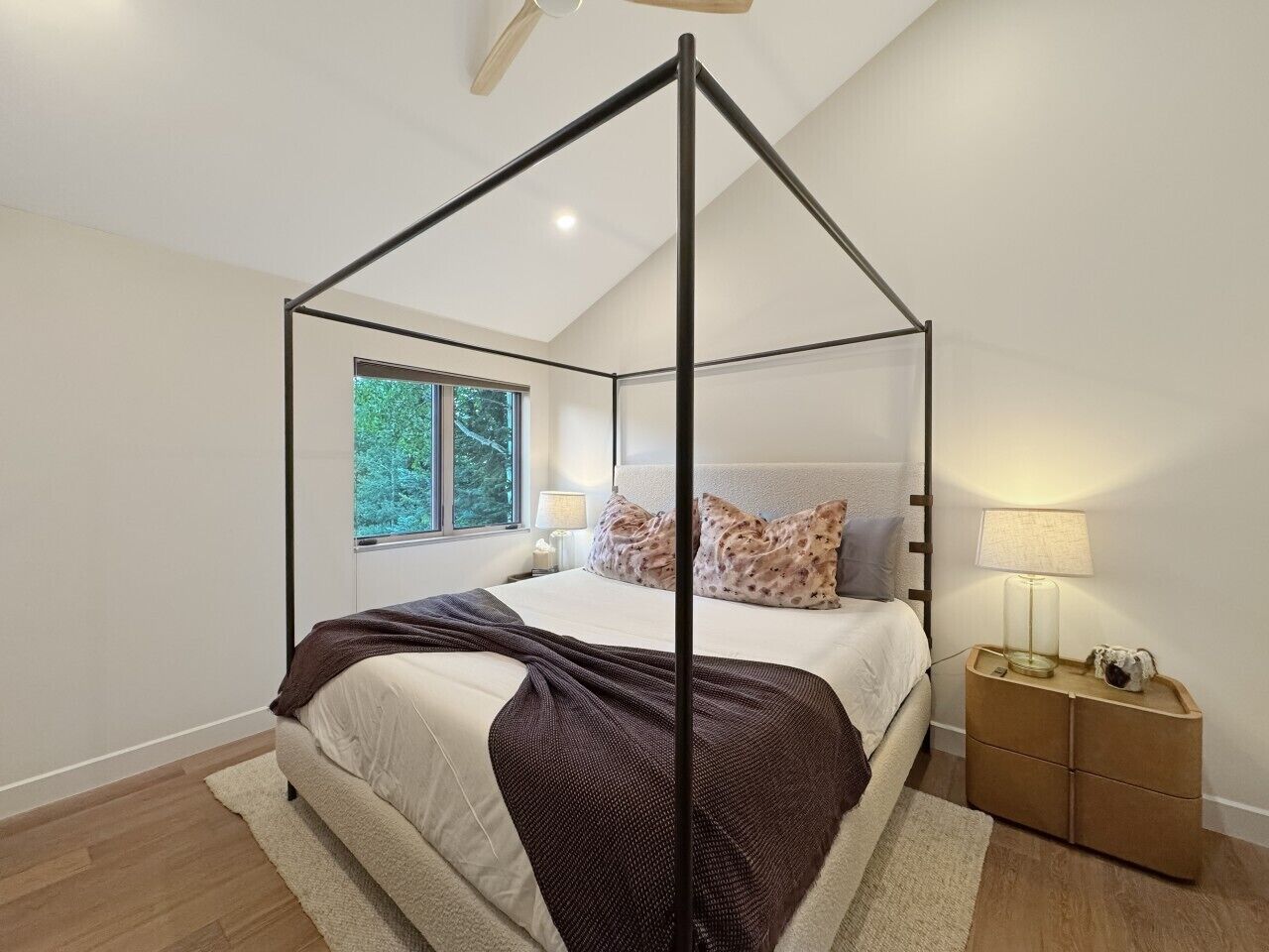 Un dormitorio con cama con dosel, una mesita de noche con lámpara y vistas a árboles a través de una ventana.