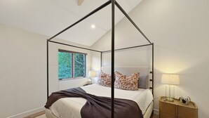 Ein Schlafzimmer mit Himmelbett, Nachttisch mit Lampe und Blick auf Bäume durch ein Fenster.