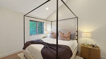 Un dormitorio con cama con dosel, una mesita de noche con lámpara y vistas a árboles a través de una ventana.