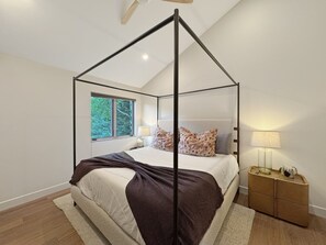 Ein Schlafzimmer mit Himmelbett, Nachttisch mit Lampe und Blick auf Bäume durch ein Fenster.