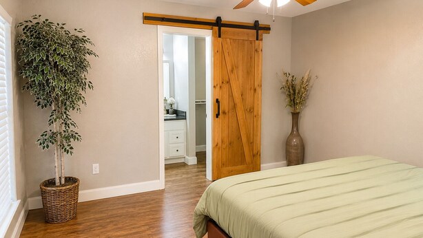 A bedroom with a wooden floor, a bed with a green bedspread, a sliding barn door, a potted plant, and a vase with dried flowers.