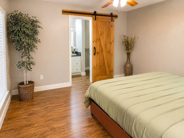 A bedroom with a wooden floor, a bed with a green bedspread, a sliding barn door, a potted plant, and a vase with dried flowers.