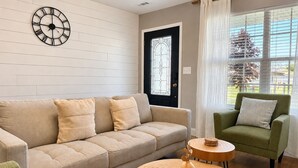A living room with a beige sofa, a green armchair, a round wooden coffee table, and a wall clock.