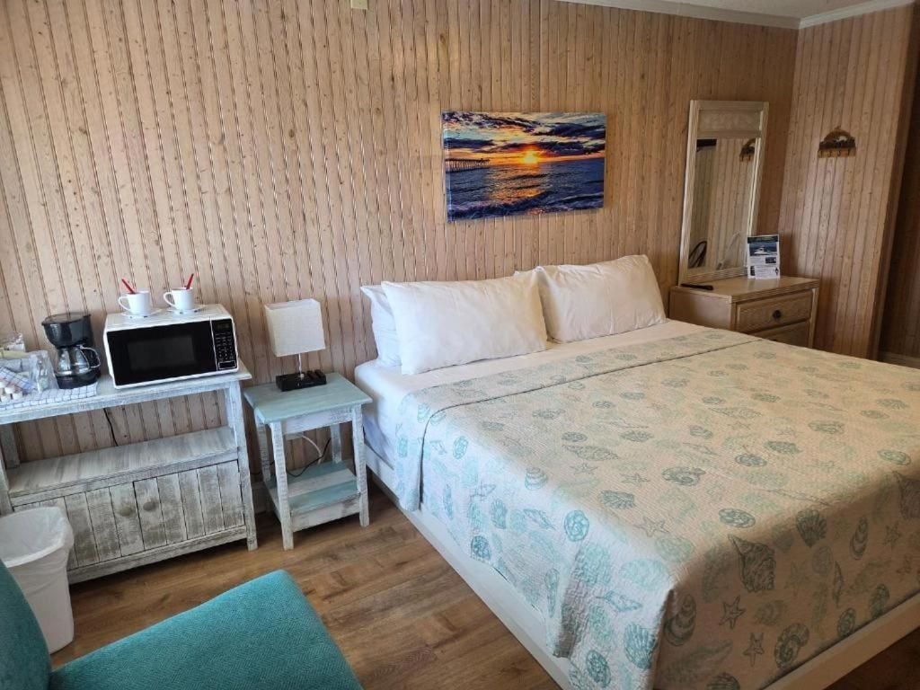 A hotel room with a bed, a desk with a microwave, a nightstand, and a painting of a sunset.