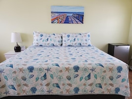Una cama con una colcha estampada, una mesita de noche con una lámpara, un cuadro en la pared y un escritorio pequeño con una computadora.