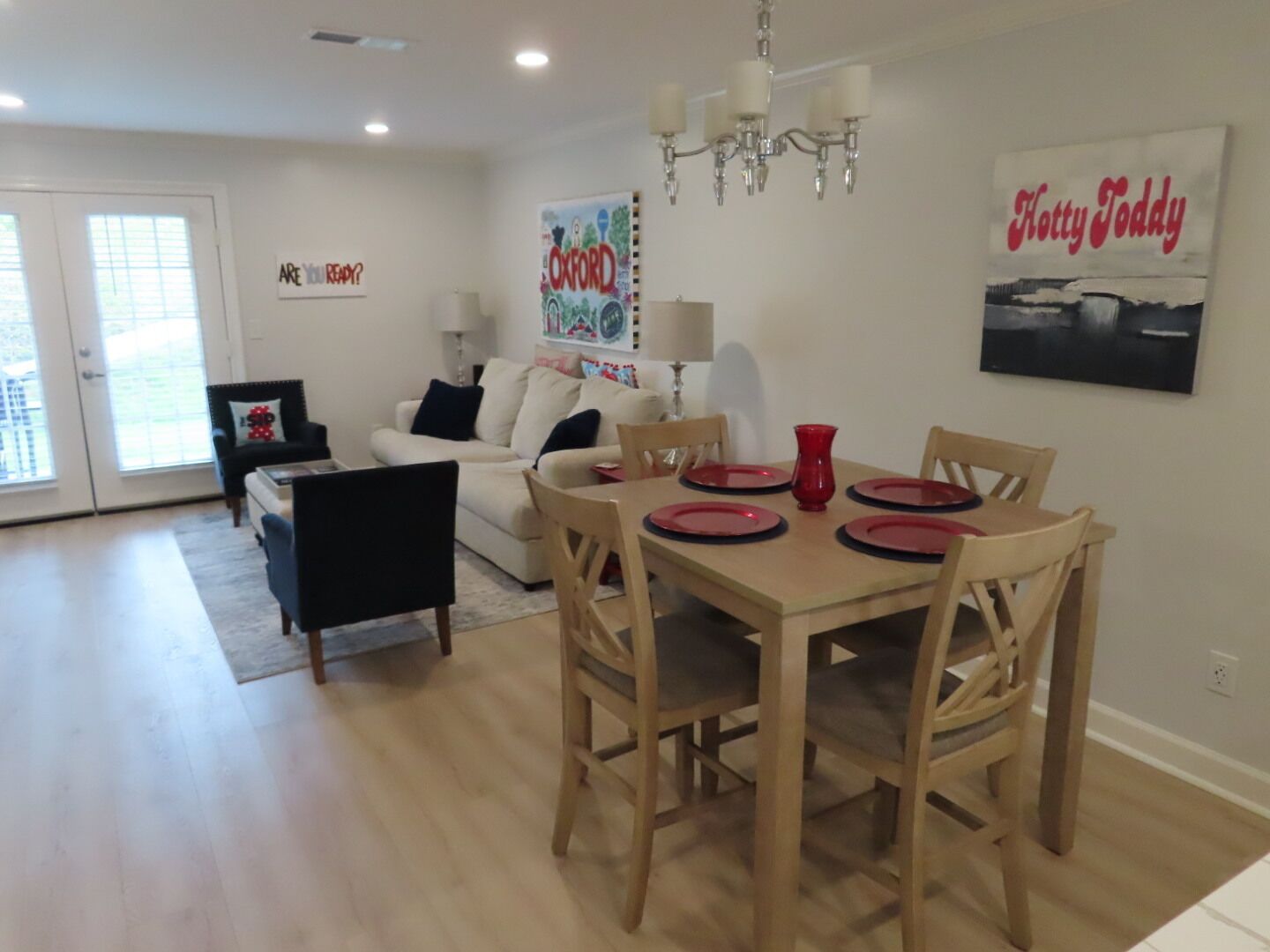 Un coin repas avec une table en bois dressée pour quatre personnes, un lustre et un tableau encadré représentant une œuvre de « Katty Todd » accroché au mur.