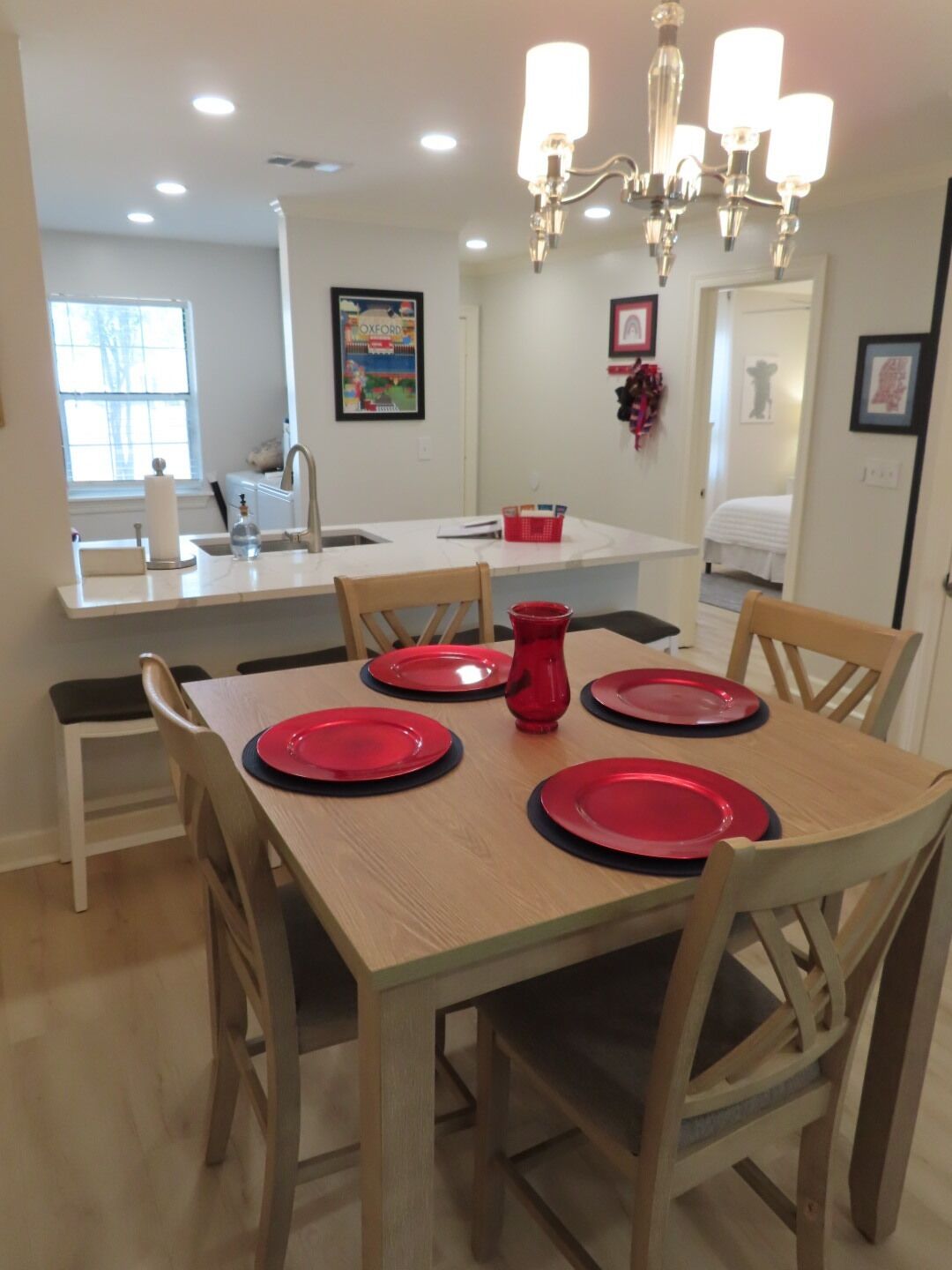 A dining area with a wooden table set for four, a chandelier, and a kitchen in the background.
