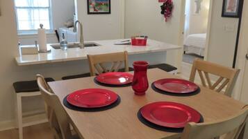 A dining area with a wooden table set for four, a chandelier, and a kitchen in the background.