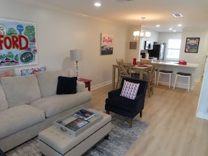 A living room with a sofa, armchair, and ottoman, a dining area with a table and chairs, and a kitchen in the background.