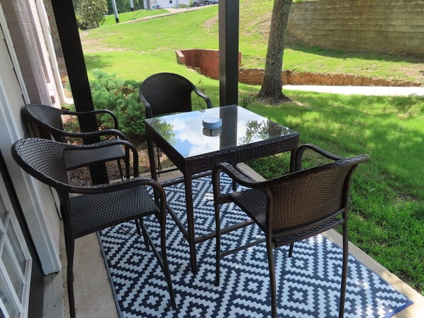 A patio area with a glass table and four wicker chairs, a patterned rug, and a view of a grassy yard.