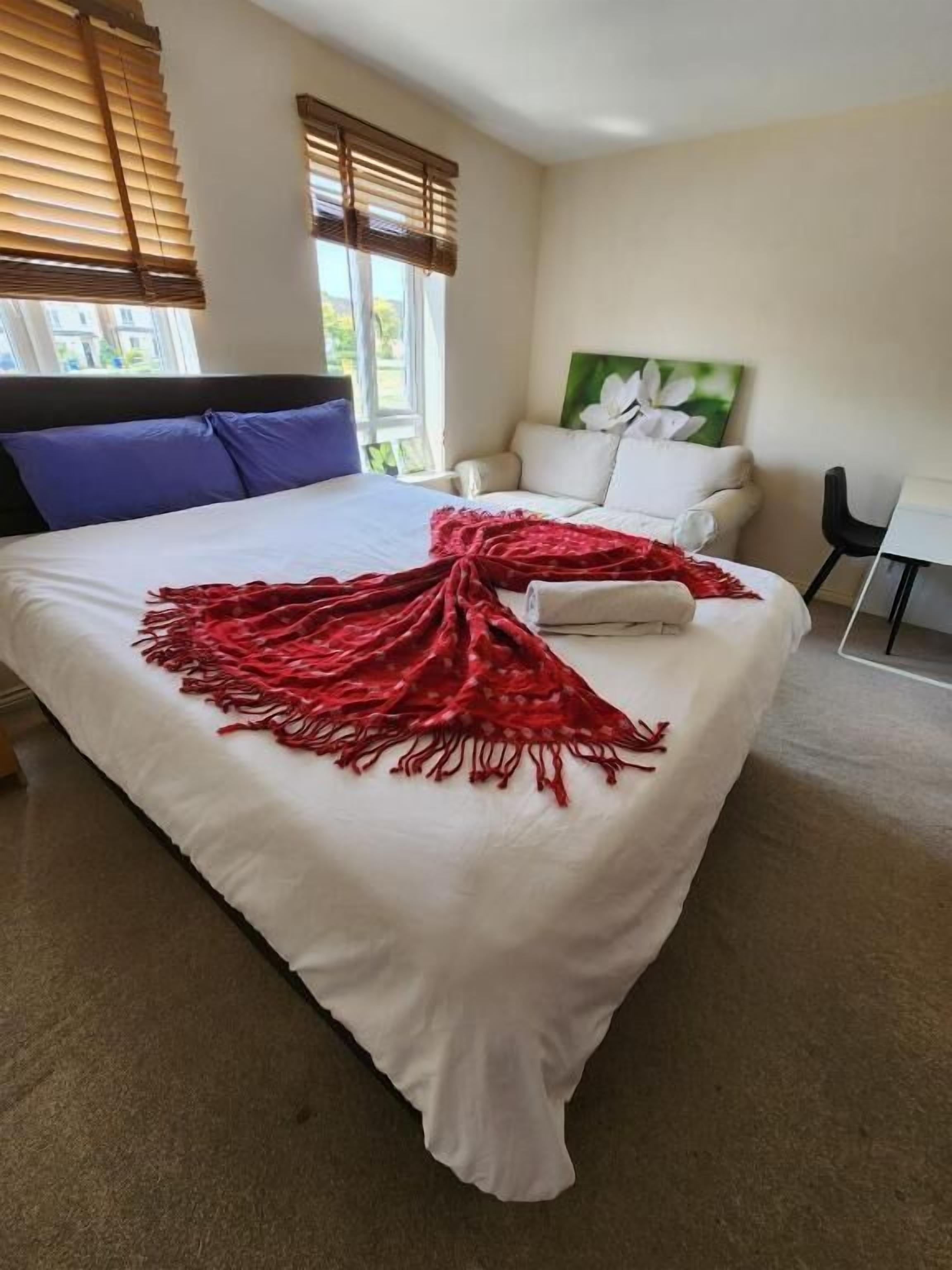 A bedroom with a large bed, a white headboard, a red throw blanket, a white sofa, and a painting on the wall.