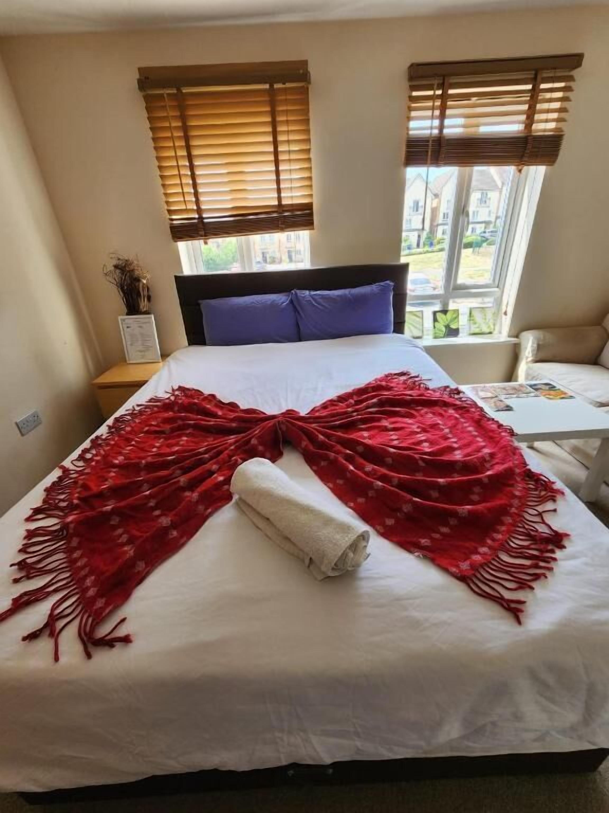 A neatly made bed with a white sheet and a red blanket, a nightstand with a lamp, and a window with blinds.