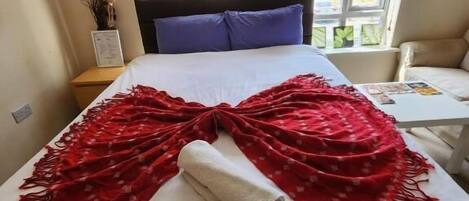 A neatly made bed with a white sheet and a red blanket, a nightstand with a lamp, and a window with blinds.