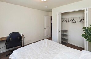 Ein Schlafzimmer mit Bett, Schreibtisch, Stuhl, Kleiderschrank mit Kleiderhaken und einer Topfpflanze.