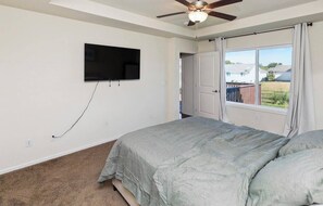A bedroom with a bed, a ceiling fan, a TV mounted on the wall, and a window with curtains. - Des Moines Ranch near Altoona | Fenced Yard, 3BR (Des Moines)
