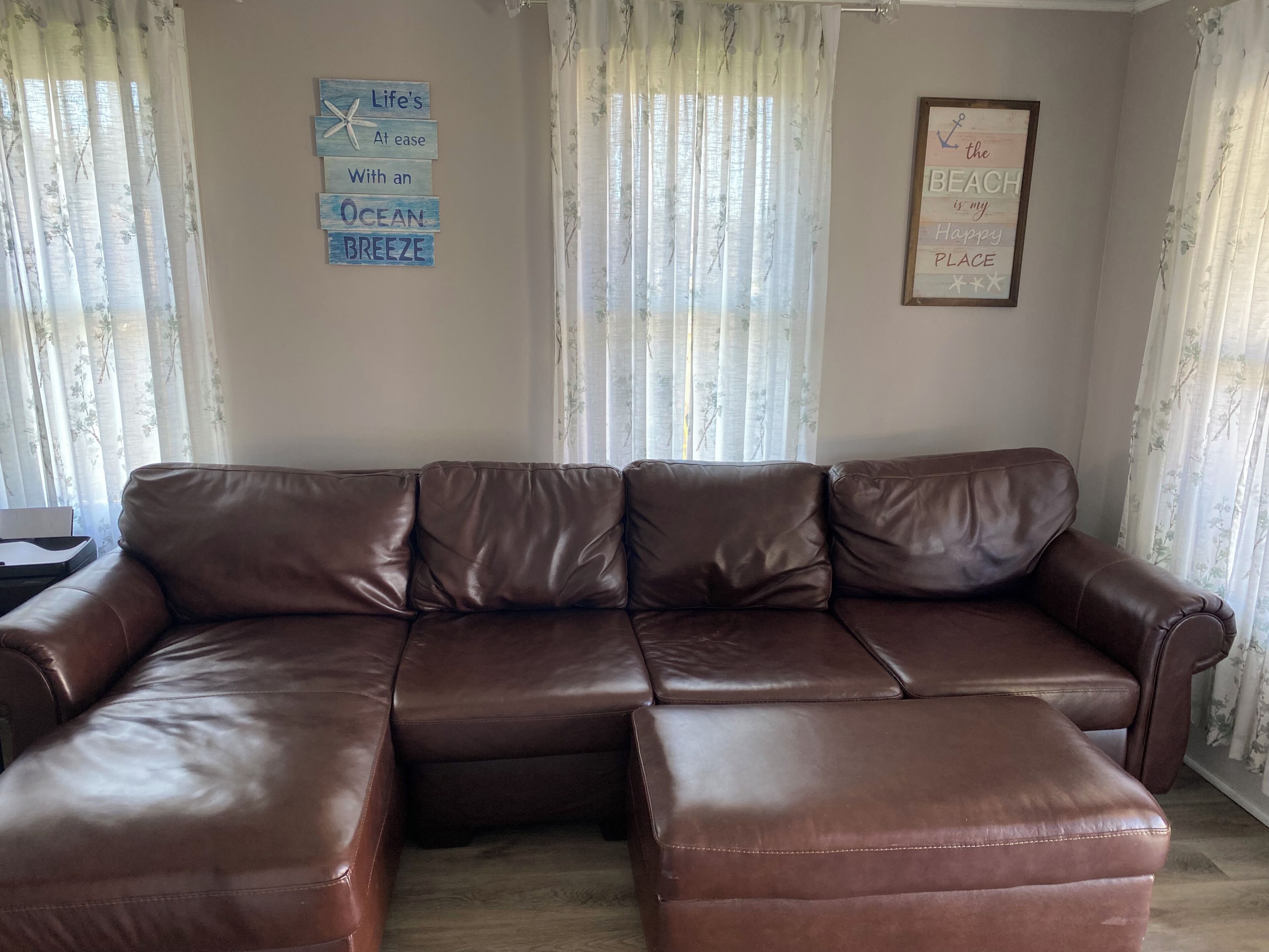 Ein braunes, ledernes Ecksofa in einem Wohnzimmer mit blumigen Vorhängen und zwei Wanddekorationen.