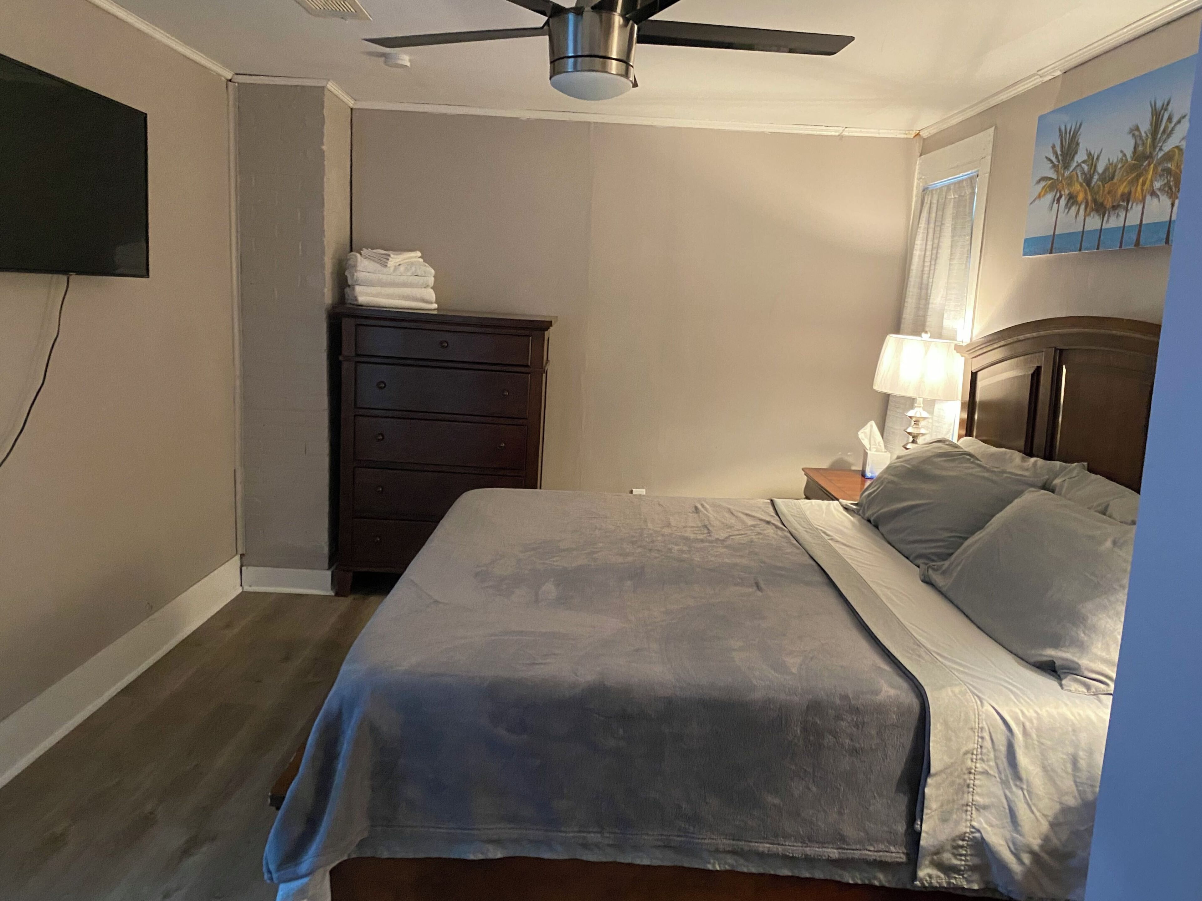 A bedroom with a bed, a dresser, a ceiling fan, a nightstand, and a wall-mounted TV.