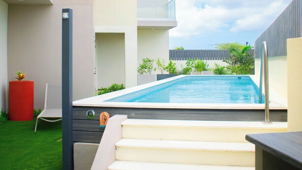 A modern outdoor swimming pool area with a clear view of the sky, a set of stairs leading to a balcony, and a small garden with a red planter. - Bonsai Boutique by BlueAruba Rentals (Noord)