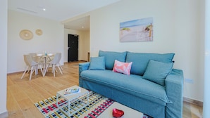 A living room with a blue sofa, a dining table with chairs, a colorful rug, and a beach-themed painting on the wall. - Bonsai Boutique by BlueAruba Rentals (Noord)