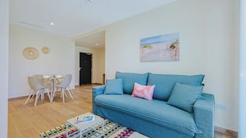 Uma sala de estar com um sofá azul, uma mesa de jantar com cadeiras, um tapete colorido e uma pintura com tema de praia na parede.