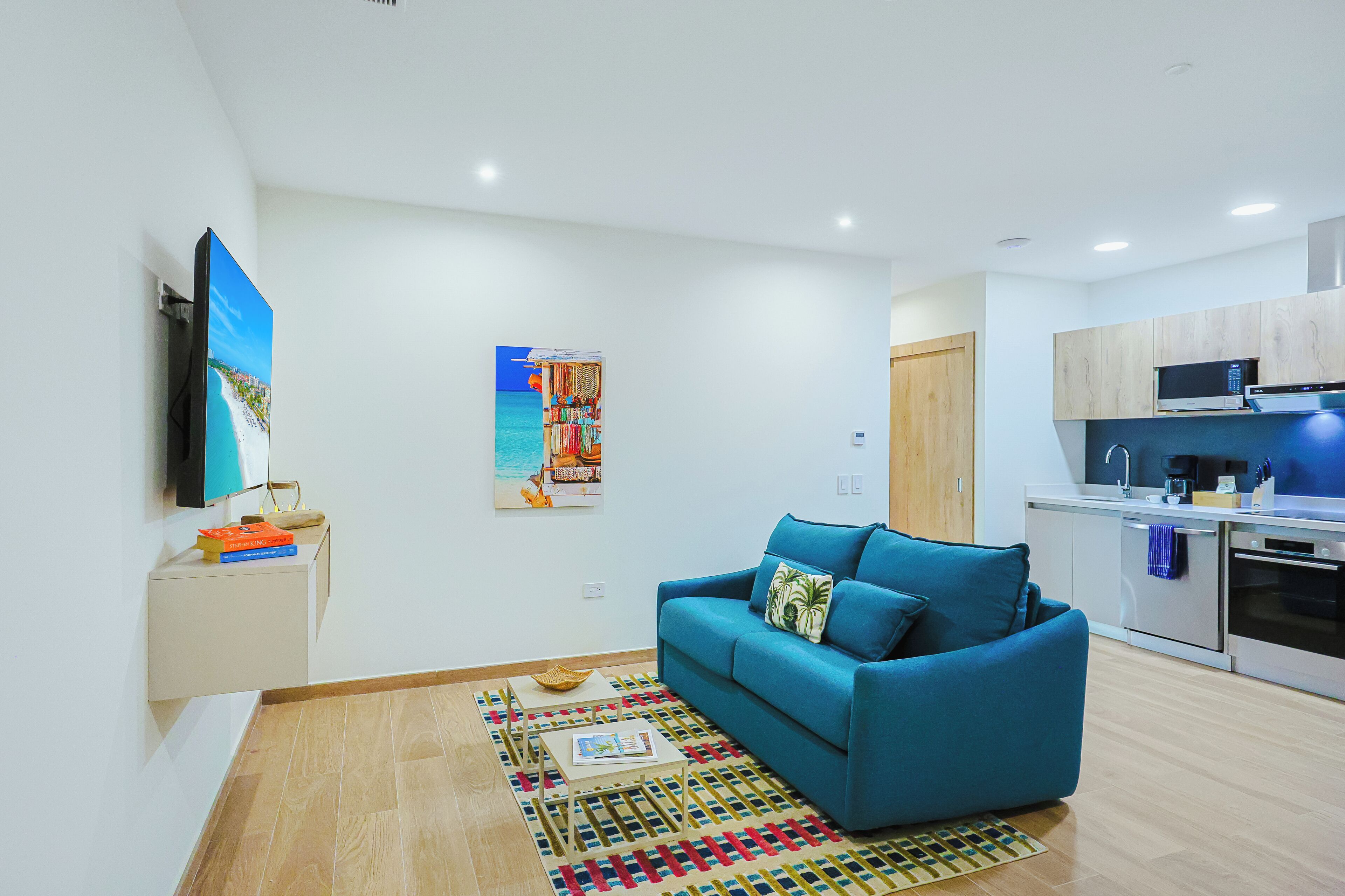 A modern living room with a blue sofa, a flat-screen TV mounted on the wall, and a kitchen area with stainless steel appliances.