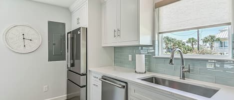 A modern kitchen with white cabinetry, a stainless steel refrigerator, a dishwasher, and a window with a view of palm trees.