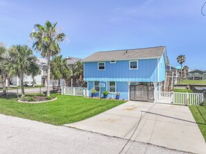 A two-story residential building with a blue exterior, a front yard with a white picket fence, and a driveway leading to the entrance. - Blue Haven | Canal-front retreat | Boat-Slip (Galveston)