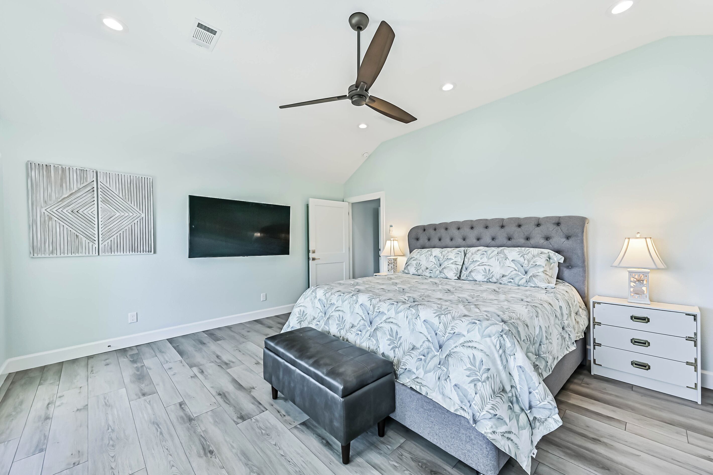 Une chambre à coucher comprenant un lit, une table de chevet, un banc, un ventilateur de plafond et des œuvres d’art accrochées aux murs.
