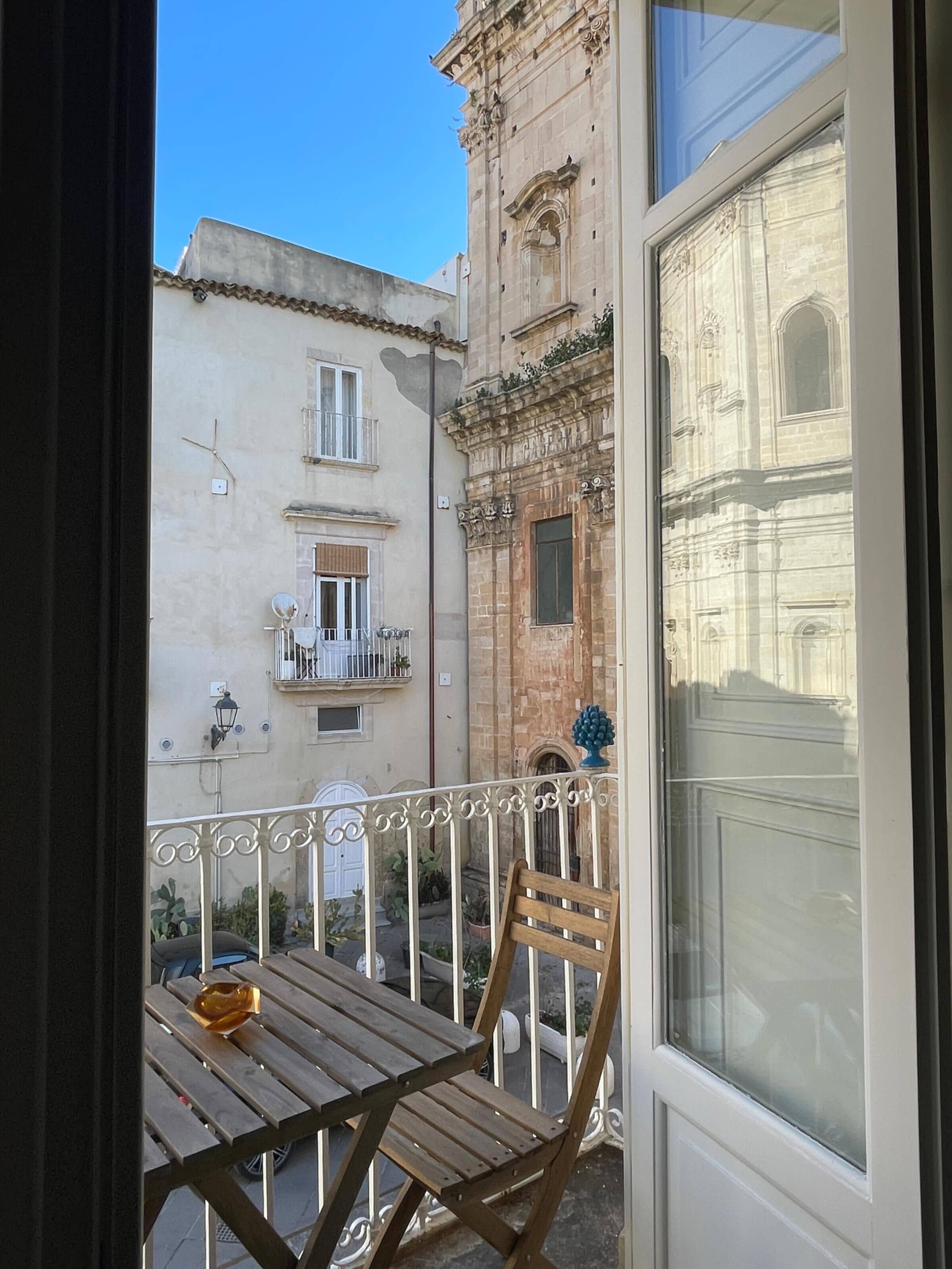 A balcony with a wooden table and chair, a small potted plant, and a view of a building with a bell tower.
