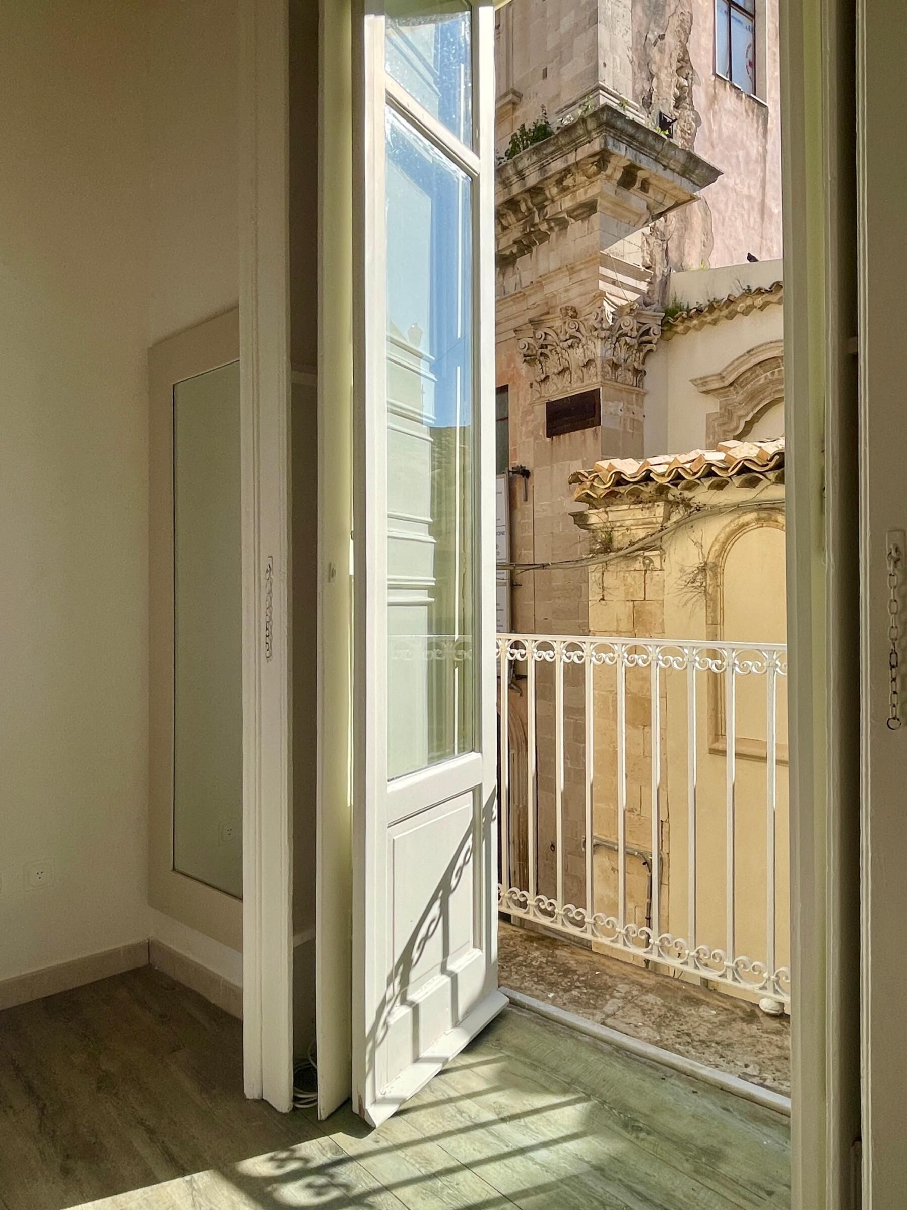Une porte ouverte donnant sur un balcon orné d’une balustrade en fer forgé, avec vue sur un immeuble aux détails architecturaux raffinés, et un plancher en bois à l’intérieur.
