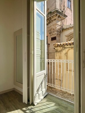Une porte ouverte donnant sur un balcon orné d’une balustrade en fer forgé, avec vue sur un immeuble aux détails architecturaux raffinés, et un plancher en bois à l’intérieur.