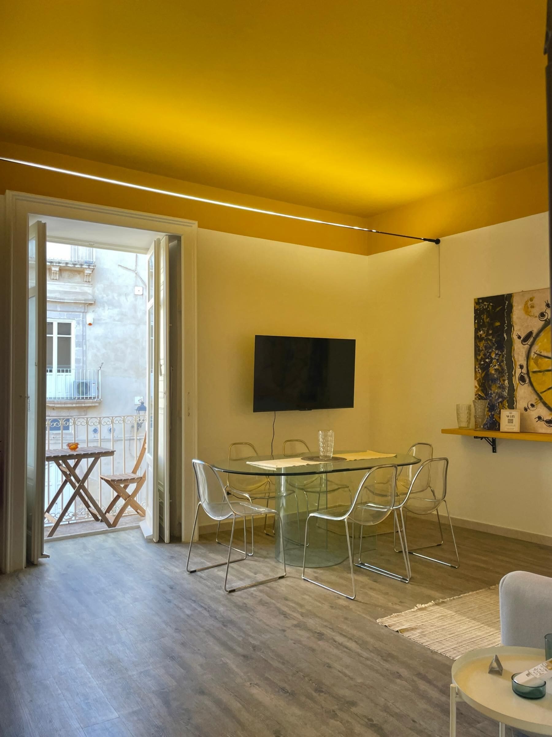 Un salon moderne avec une table à manger en verre, des chaises transparentes, un téléviseur à écran plat fixé au mur et un balcon offrant une vue sur un immeuble.