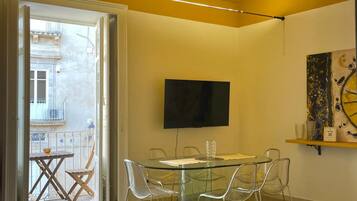 A modern living room with a glass dining table, clear chairs, a flat-screen TV mounted on the wall, and a balcony with a view of a building.