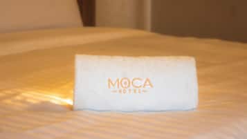 Una cama bien tendida con una toalla doblada encima, mostrando el texto 'MOCA HOTEL'.
