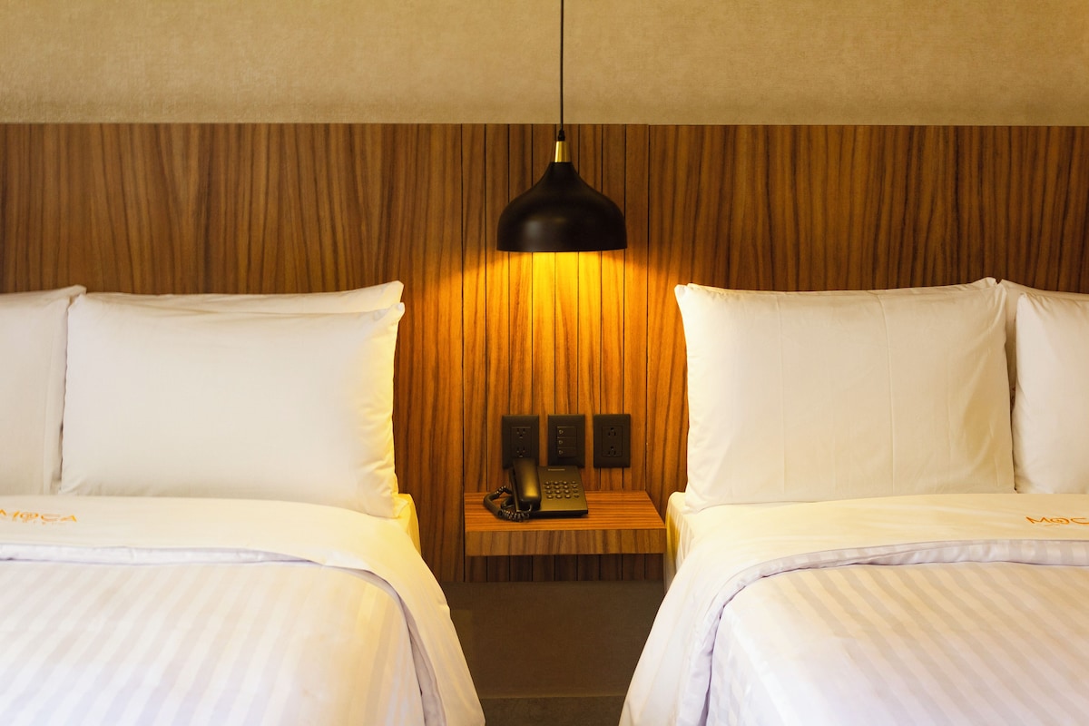 A hotel room with two beds, a wooden headboard, a bedside table with a phone and a lamp, and a wall-mounted power outlet.