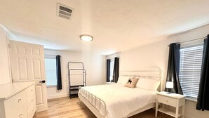Een slaapkamer met een bed, nachtkastje, lamp, kast en een raam met gordijnen.