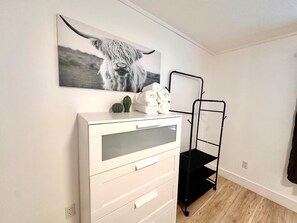 Un comò bianco con cassetti, una scaffalatura in metallo nero, un quadro con uno yak e un piccolo cactus sul comò.