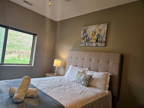 Ein Schlafzimmer mit einem großen Bett, einem Nachttisch mit Lampe, einem Fenster mit Blick ins Grüne und einem Gemälde an der Wand.