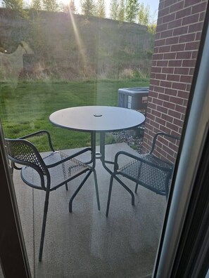 A patio with a round table and four chairs, a brick wall, and a grassy area in the background. - 1-bed by Jordan Creek & Top Golf (West Des Moines)