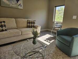A living room with a sofa, a chair, a coffee table, and a window with a view of greenery. - 1-bed by Jordan Creek & Top Golf (West Des Moines)