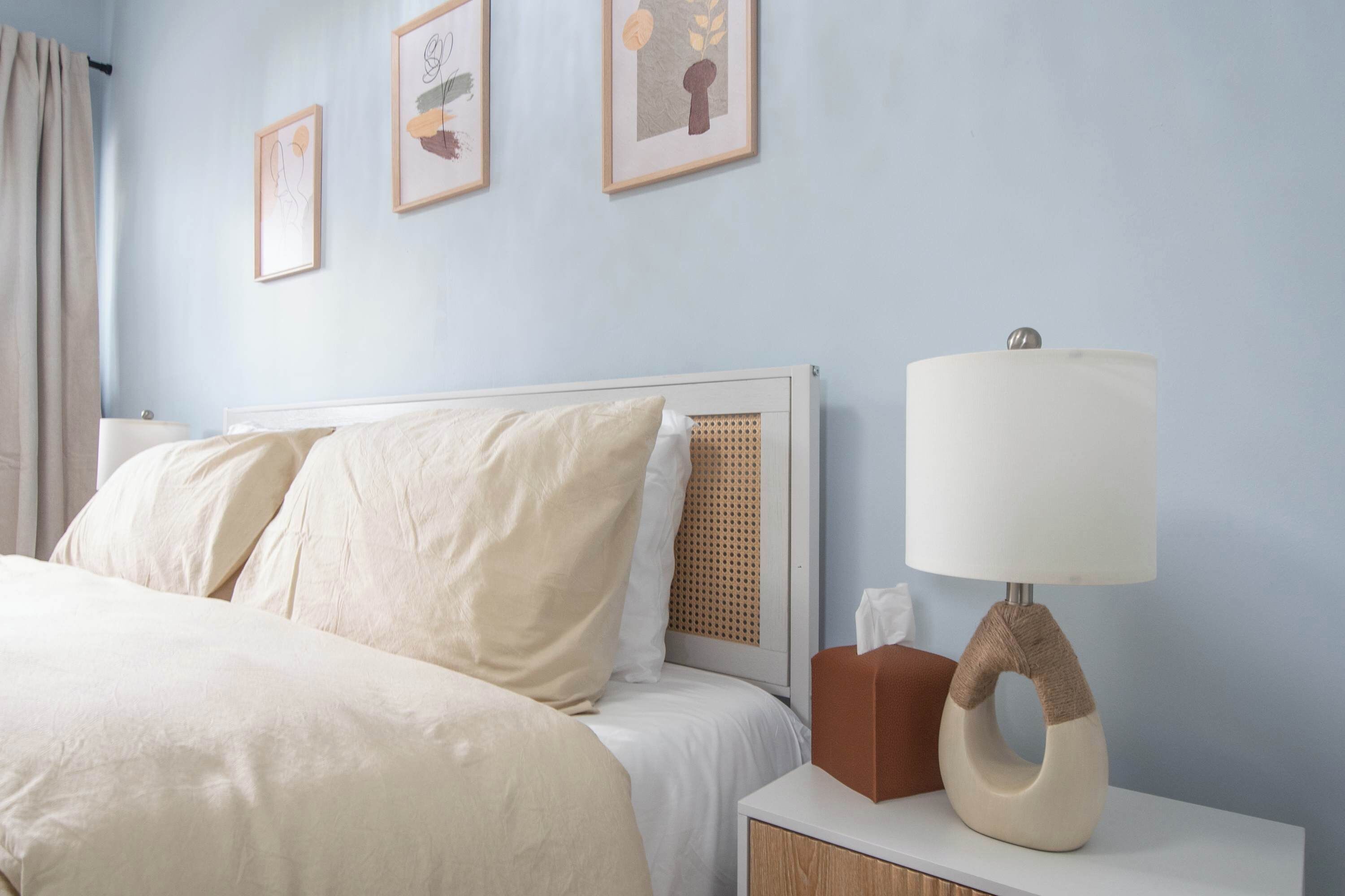Un lit bien fait, avec une tête de lit blanche et deux oreillers, une table de chevet avec une lampe et un objet décoratif, et trois œuvres d’art encadrées accrochées au mur.