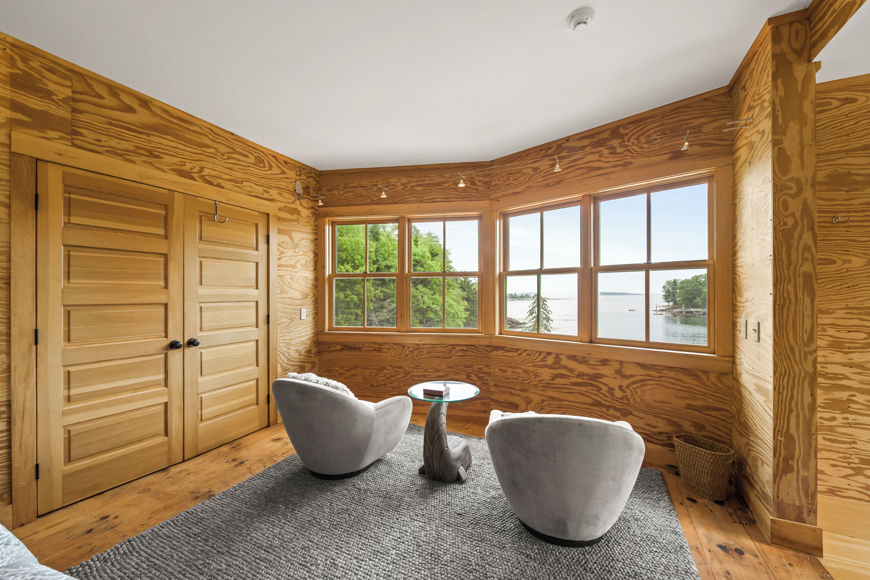 A room with wooden walls, a large window with a view of trees and water, two grey armchairs, and a small round table.