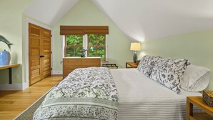 Ein Schlafzimmer mit schrägen Wänden, einem Bett mit Blumenmuster auf der Bettwäsche, einer Holzkommode und einem Fenster mit Blick auf Grünflächen.