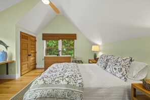 A bedroom with a sloped ceiling, a bed with floral bedding, a wooden dresser, and a window with a view of greenery. - NEW! Quaint 2BR apt in Harpswell with private beach area and beautiful gardens (Harpswell)