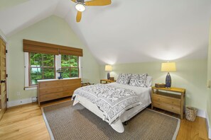 A bedroom with a sloped ceiling, a bed with a floral bedspread, two bedside lamps, a window with blinds, and a wooden dresser. - NEW! Quaint 2BR apt in Harpswell with private beach area and beautiful gardens (Harpswell)