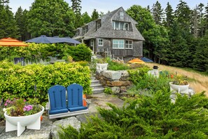 Une maison avec un escalier en pierre menant à une terrasse en bois, entourée d’une végétation luxuriante et dotée d’un espace salon extérieur.